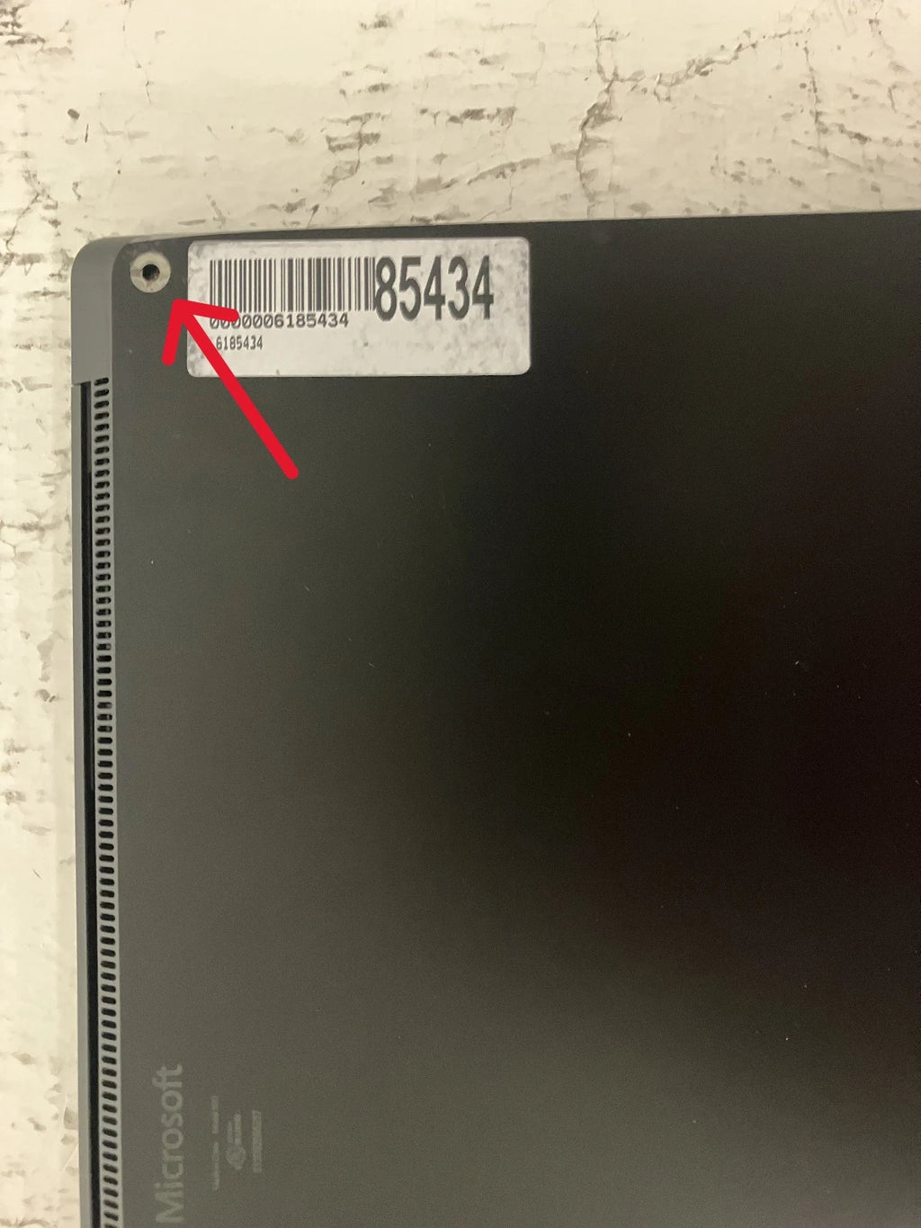 Back view of a Microsoft product with a barcode label on a textured surface and section indicating where the rubber foot is missing with a red arrow drawn on screen.